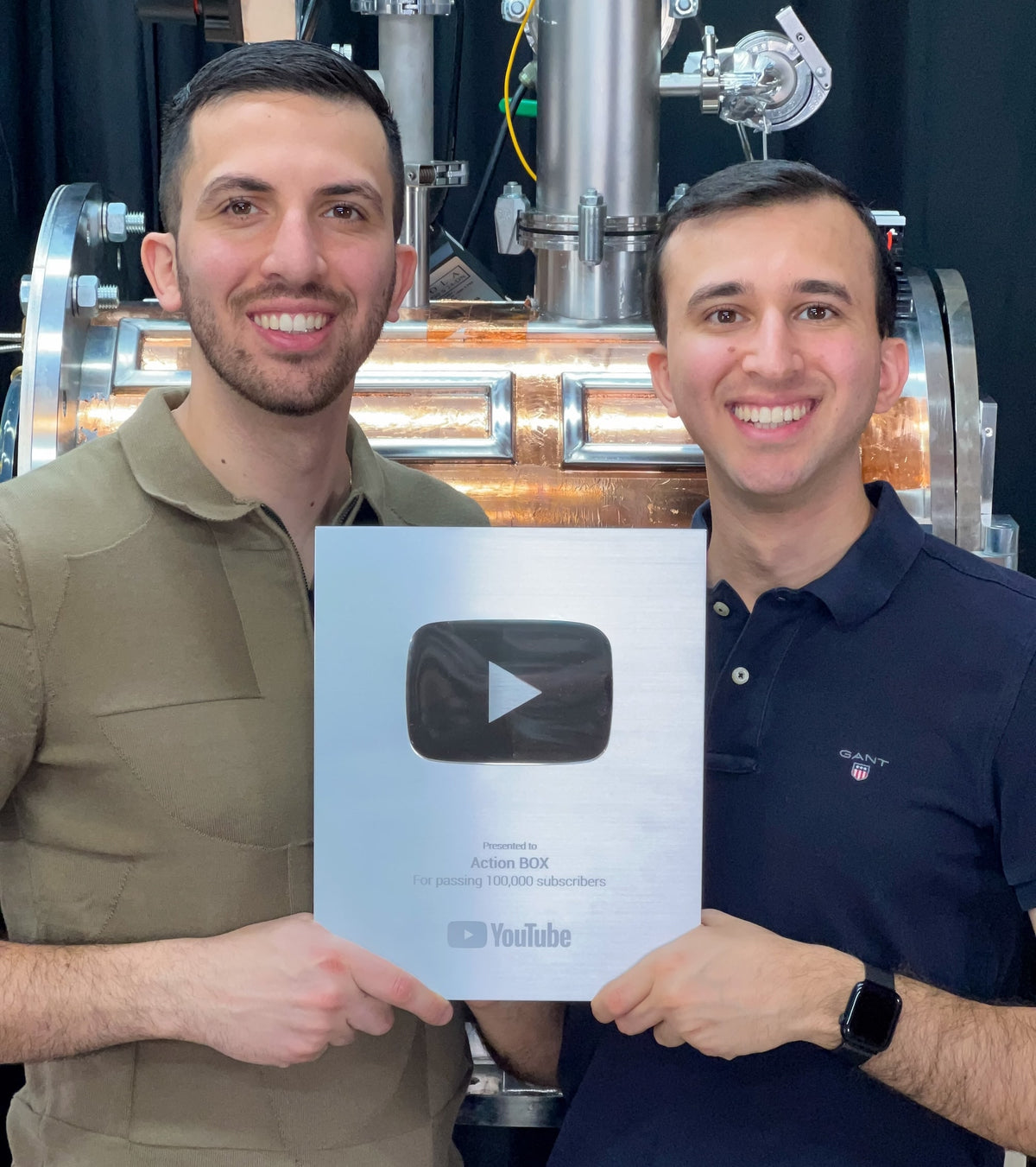 Two founders of Action BOX holding a YouTube award in front of scientific equipment they developed.