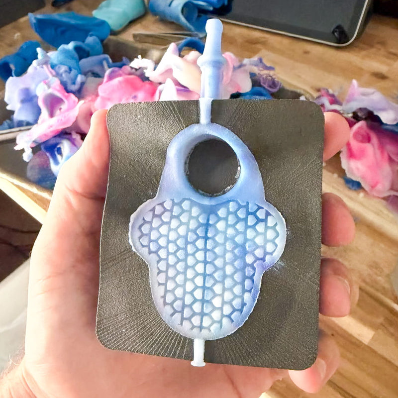A hand holds a 3D-printed mold with a light blue and white textured Hit Hero prototype, set against a workshop background with colorful injection molded TPE prototypes of Hit Hero using INJEKTO 3.